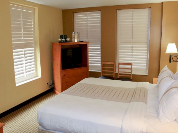 A tidy hotel room with two queen beds, a wooden dresser with a TV, and large windows with white blinds—bright and relaxing.
