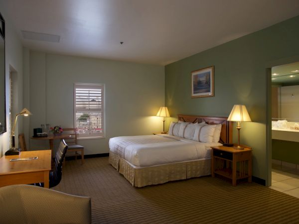 A hotel room with two double beds, green accent wall, nightstands, lamps, a desk, chair, and a doorway to a bathroom; calm, well-lit space.