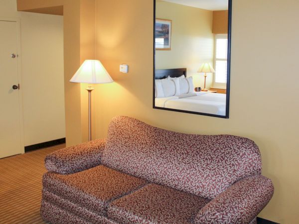 A hotel room scene with a patterned couch, a floor lamp, a large wall mirror, and a doorway; the bed and other furnishings appear in the reflected view, bright and tidy.