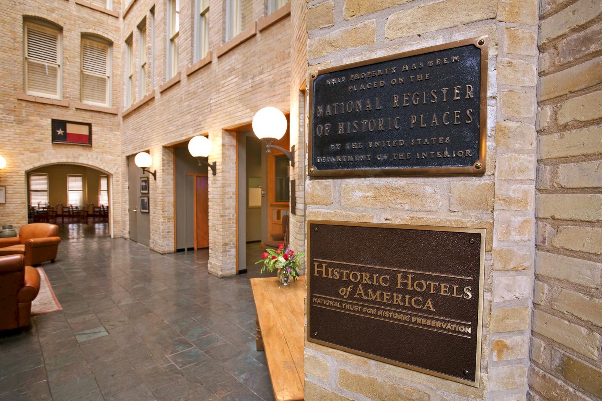 A historic hotel lobby with brick walls, hanging lamps, and plaques reading “National Register of Historic Places” and “Historic Hotels of America.”