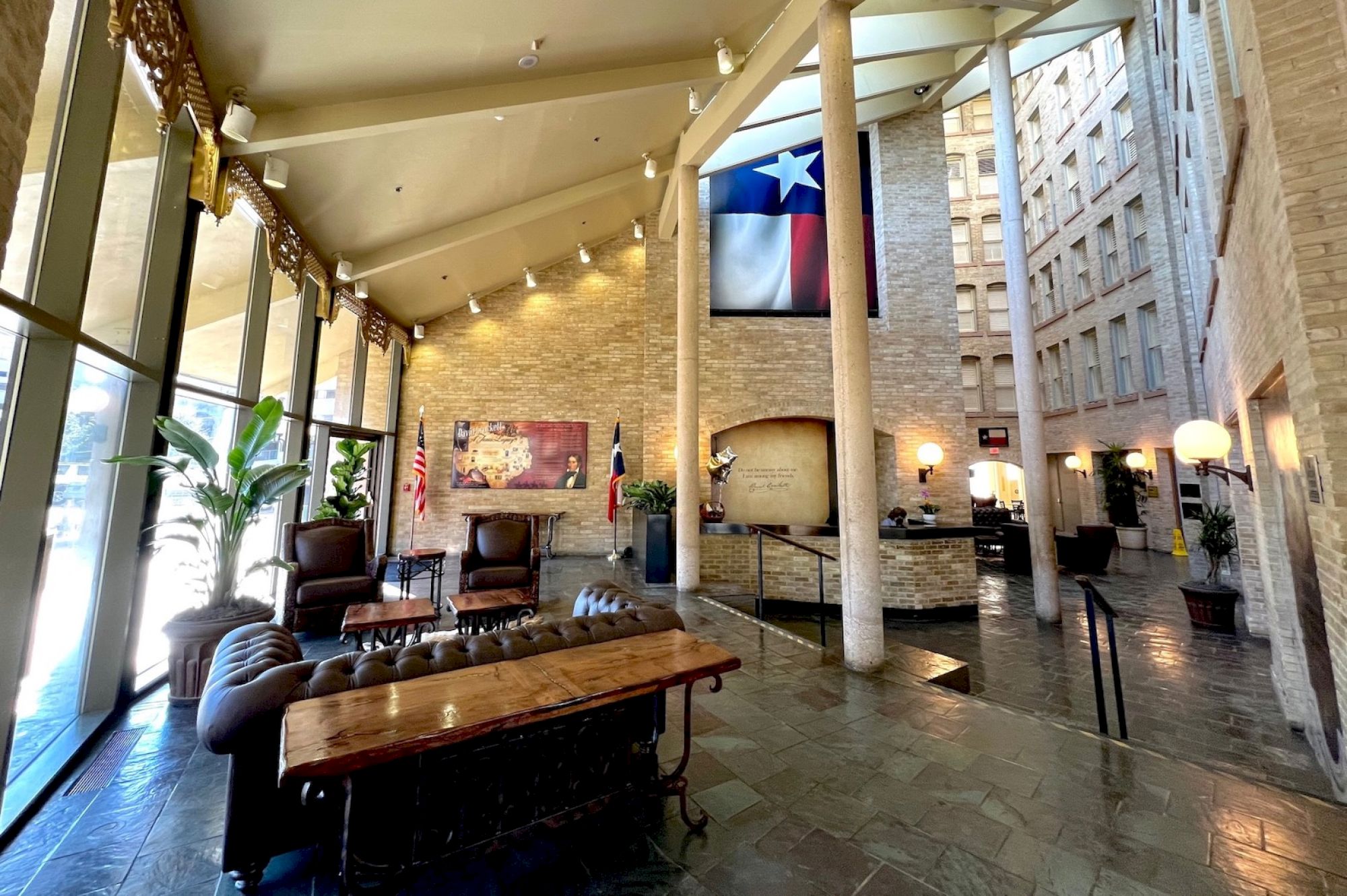A spacious hotel lobby with tall ceilings, stone floors, and comfortable seating; a Texas flag hangs from a second-floor balcony, warm lighting, and trees and artwork add to the inviting atmosphere.