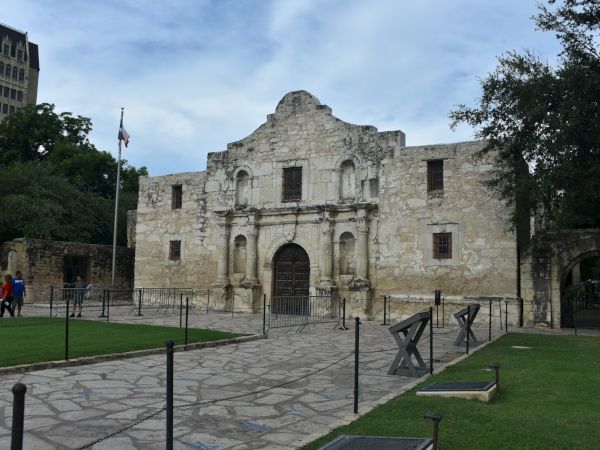 A historic stone mission-style building with an arched entrance, front courtyard, and surrounding lawn, under a blue sky.