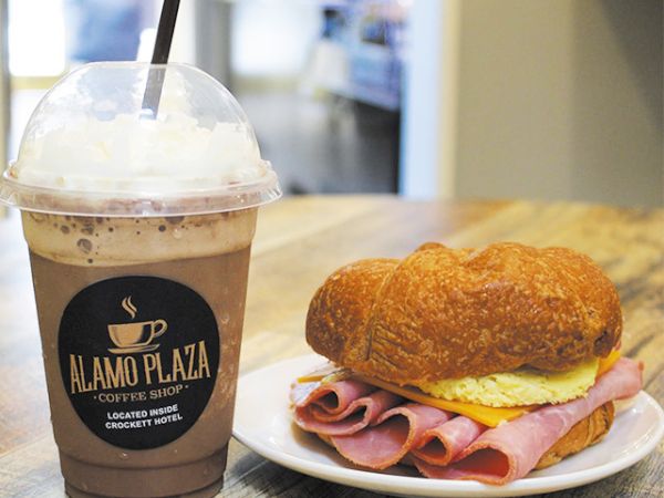 A chocolate frappe with whipped cream beside a ham and egg croissant sandwich on a table at a cafe.