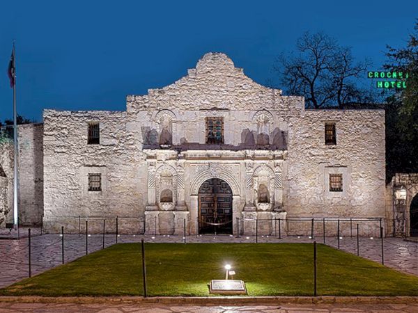 Ancient stone mission-style facade with arched doorway, flanked by small windows, a manicured lawn, and a lit pedestal in front at dusk.