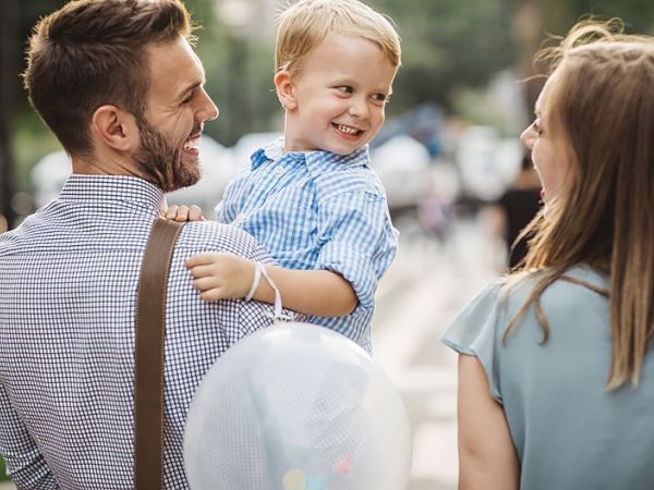 A happy family: a man with a baby boy on his shoulder, smiling at a woman; outdoor setting with balloons in the background.