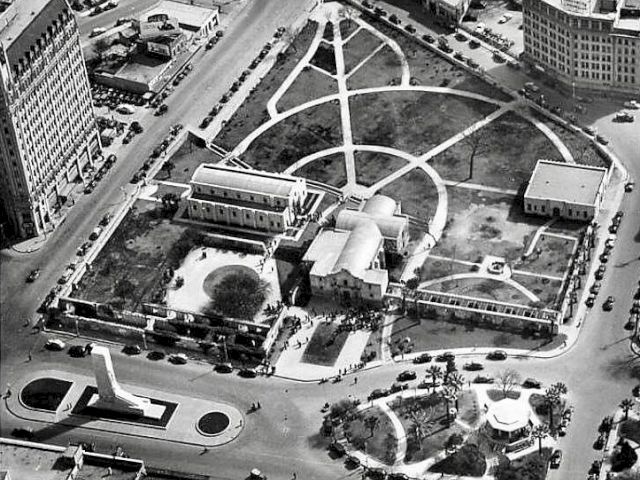 An aerial view of a curved, geometric park with paths, trees, and surrounding buildings; a central pavilion area and circular features.