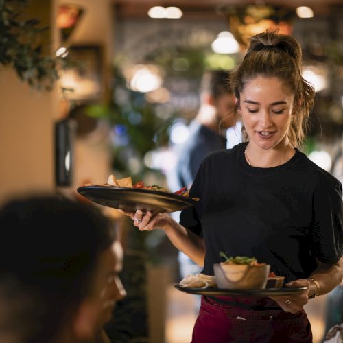 A smiling waitress carrying two trays with dishes in a busy restaurant.