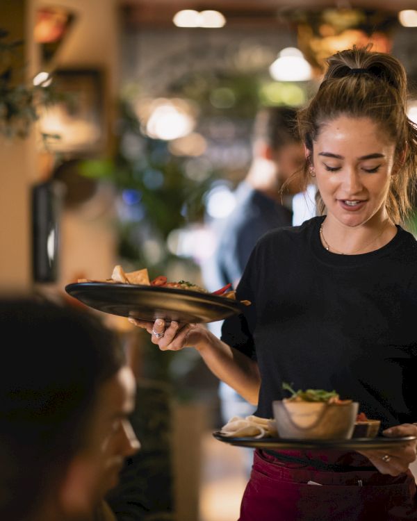 A smiling waitress carrying two trays with dishes in a busy restaurant.