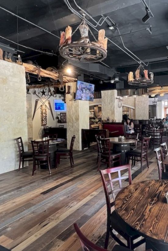 A rustic pub-style dining area with wooden tables and chairs, warm lighting, exposed brick and walls, and chandeliers hanging from a dark ceiling.
