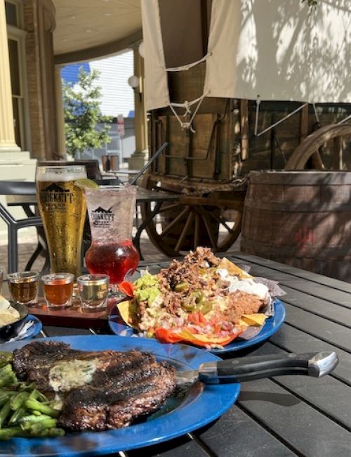 A rustic outdoor meal: two plates with steak, greens, and sides on a dark table, plus drinks (beer, water, and a red beverage) and a wagon in the background, under a shaded porch. end with a period.