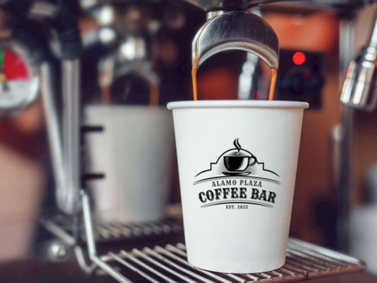 A white coffee cup with “Alamo Plaza Coffee Bar” logo sits under a coffee machine nozzle, ready to brew, against metal equipment.
