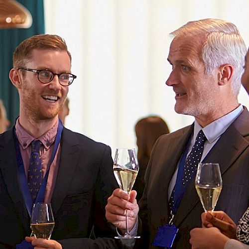 A group of professionals in business attire mingle at a social event, holding drinks and chatting warmly in a bright, modern setting.