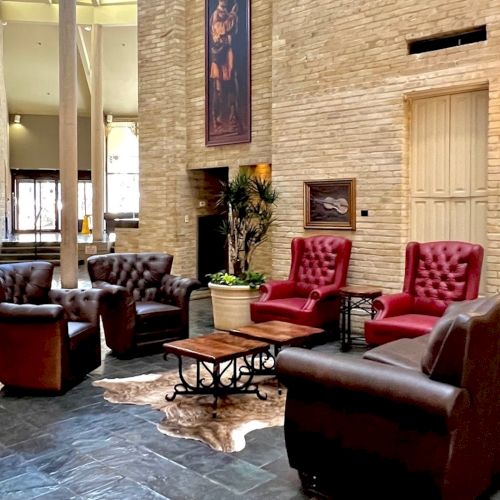 A hotel lobby with leather sofas arranged around a coffee table, beige brick walls, framed art, potted plants, and warm wall lamps. End.