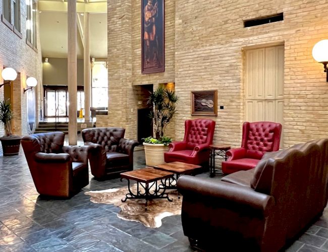 A hotel lobby with leather sofas arranged around a coffee table, beige brick walls, framed art, potted plants, and warm wall lamps. End.