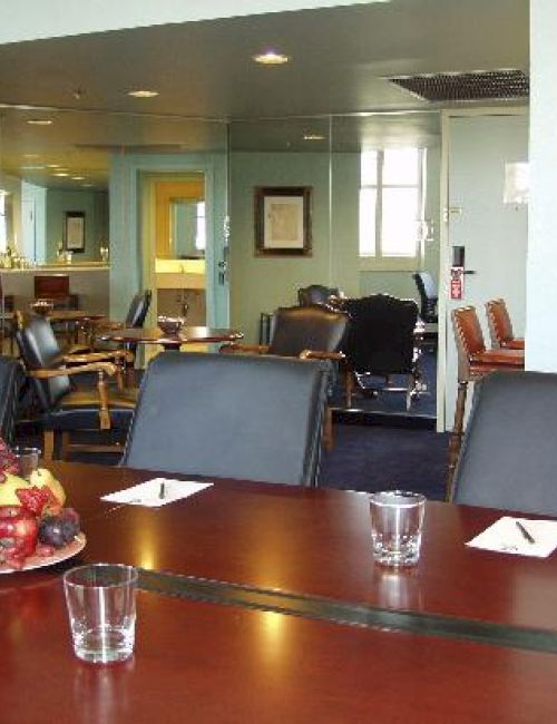 A conference room with a long wooden table, fruit centerpiece, water glasses, and padded chairs; more seating and windows visible in the background.