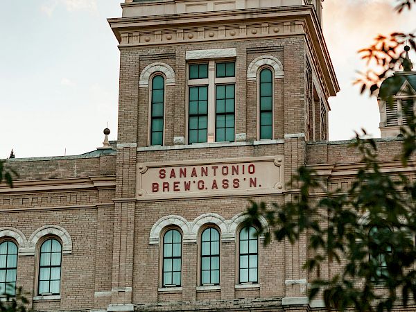 A tall historic brick building with a green domed roof and arched windows, labeled “San Antonio Brev. Kass.” at the facade.