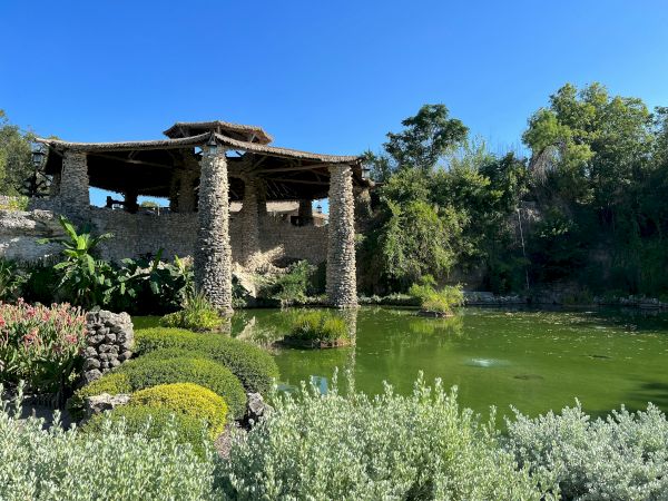 A stone gazebo over a bright green pond, surrounded by lush shrubs and trees on a sunny day.
