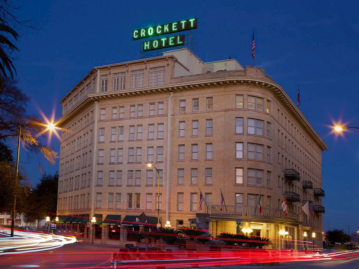 A historic brick hotel at dusk with “Crockett Hotel” sign on top, street lights, and light trails from passing cars.