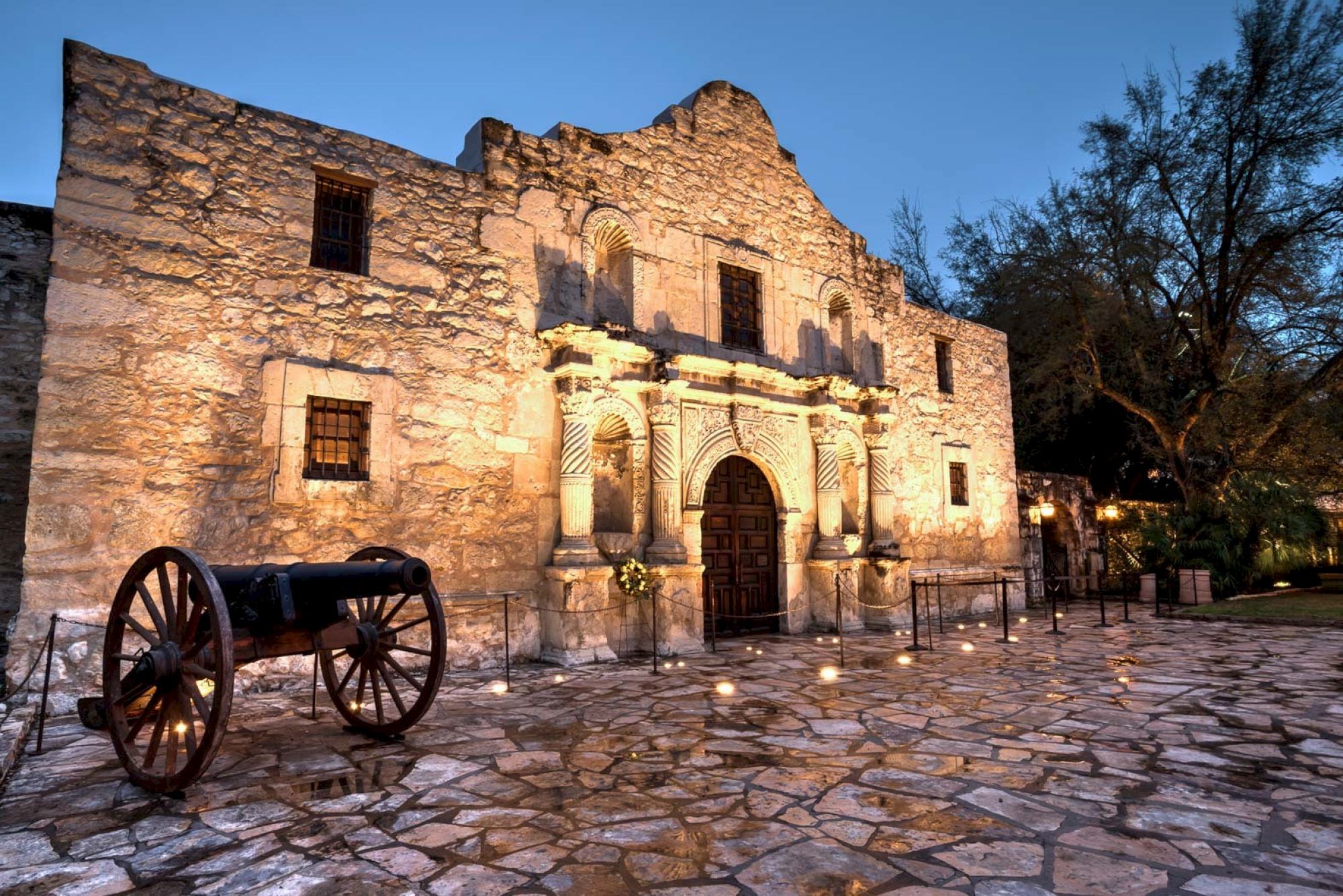 Ancient stone fortress with arched doorway, illuminated facade, rustic cannon on wheels, and cobblestone courtyard at twilight.