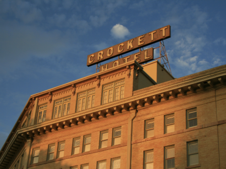 A brick hotel with a large rooftop sign reading “CROCKETT HOTEL” against a blue sky with a flag on a pole atop the building.
