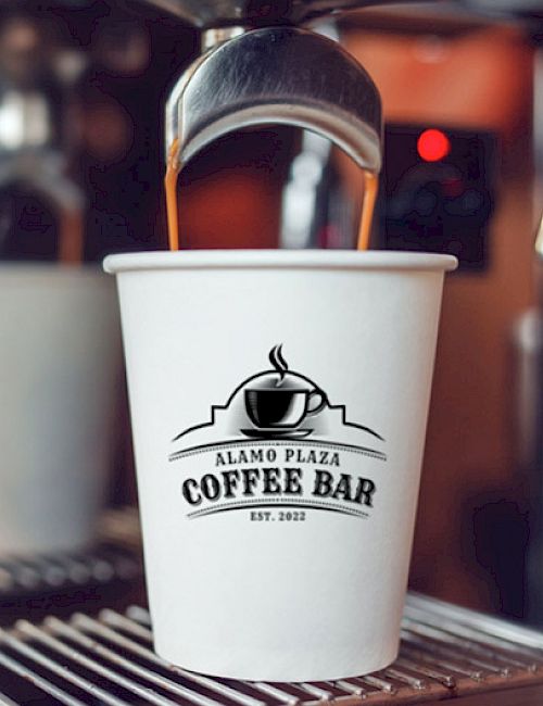 A white coffee cup with the Alamo Plaza Coffee Bar logo sits on a espresso machine, dispensed by a metal nozzle in a cozy cafe ambiance.