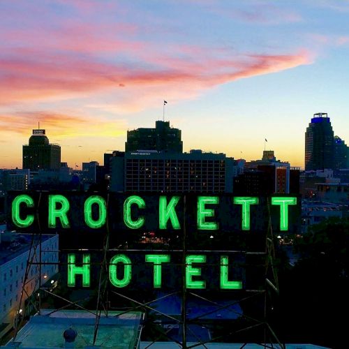 A city skyline at sunset with a neon green “CROCKETT HOTEL” sign perched above buildings, glowing against pink-orange sky and fading blue.