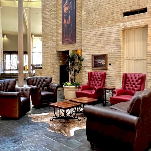 A stylish hotel lobby with brown and red leather seating around a circular coffee table, brick walls, plants, and warm lighting, inviting and cozy.
