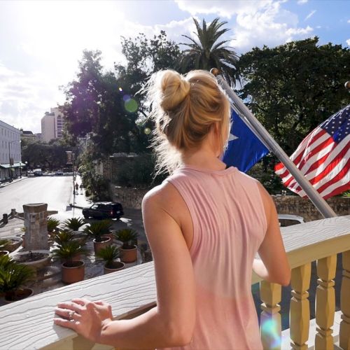 A woman in a pink sleeveless top stands on a balcony overlooking a sunny city street with potted plants and American flags waving, back to camera.