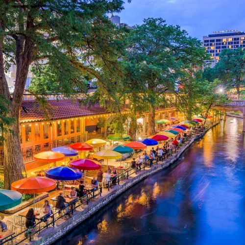 A vibrant riverside scene with a quaint walkway, colorful umbrellas shading tables, trees lining the canal, and warm lights reflecting off the water.