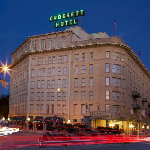 Crockett Hotel building at dusk with lights on, sign top, street traffic trails in front.