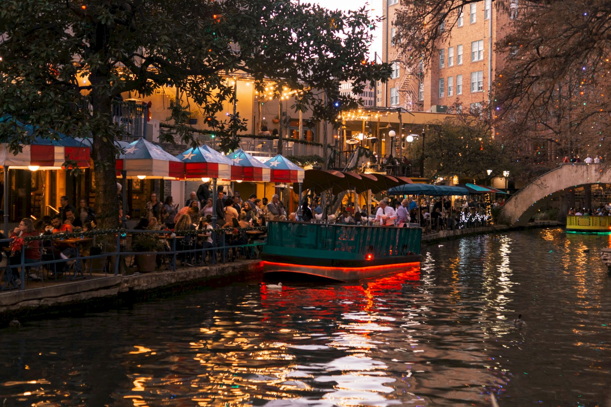 People dining along a riverfront at dusk, with a lit boat cruising past and colorful market tents lining the water.